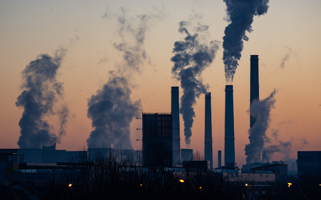 A imagem mostra uma foto de uma indústria liberando no ar gases poluentes. A foto foi tirada no crepúsculo e está quase escura. A fumaça é cinza e preta.