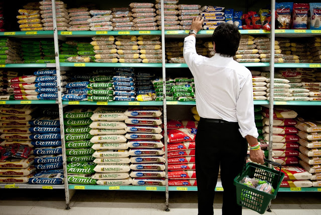 Homem de costas para a câmera escolhendo um pacote de feijão em uma prateleira de supermercado com várias marcas de arroz e feijão diferentes