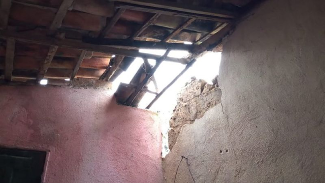 A imagem mostra uma foto de um telhado com telhas quebradas e uma parte da parede faltando devido aos tremores de terra. É uma casa com paredes rosas e brancas, as telhas são marrons e há uma estrutura de madeira por baixo.