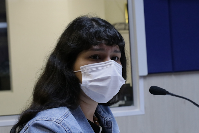A imagem mostra Renata usando máscara e uma jaqueta jeans. Ela está de lado em frente a um microfone.