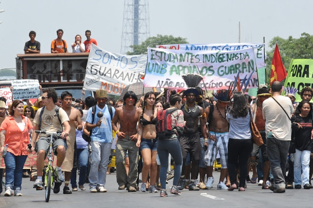 Foto de uma manifestação com estudantes e índios caminhando pela rua, de bicicleta e em cima de um trio elétrico. Algumas pessoas seguram faixas. Numa delas está escrito “somos todos Guarani” e em outra “Estudantes indígenas da UNB apoia os Guarani!”ente do nariz pra cima.