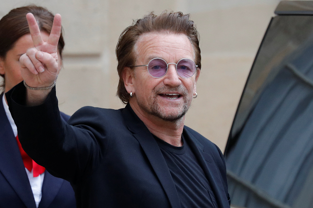 A imagem mostra o cantor Bono Vox acenando para a câmera. Ele é caucasiano, usa terno preto e um óculos roxo. Ele está sorrindo.