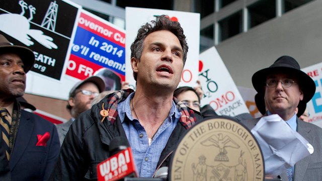 A imagem mostra o ator Mark Ruffalo em um protesto ambiental. Ele é caucasiano e veste uma jaqueta marrom, ele segura cartazes contra o fraturamento hidraúlico.