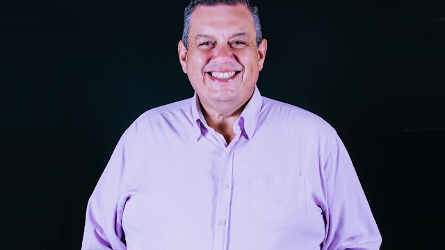 A imagem mostra Sérgio Alba sorrindo, ele veste uma camisa lisa lilás. Ele está em um fundo preto. As mãos estão no bolso.