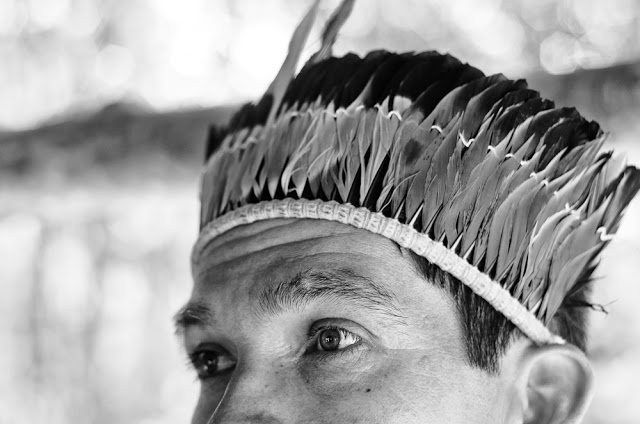Foto em preto em branco de um índio Kaiowá. O índio está com um cocar e olhando ao longe. A foto foca somente do nariz pra cima.