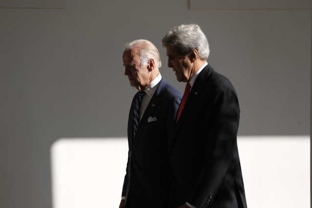 A imagem mostra Joe Biden e John Kerry andando lado a lado. A foto foi tirada da lateral dos homens. Eles são caucasianos. Joe usa um terno azul e gravata listrada com azul e branco. E John usa um terno preto com gravata vermelha. Ambos tem cabelos brancos.