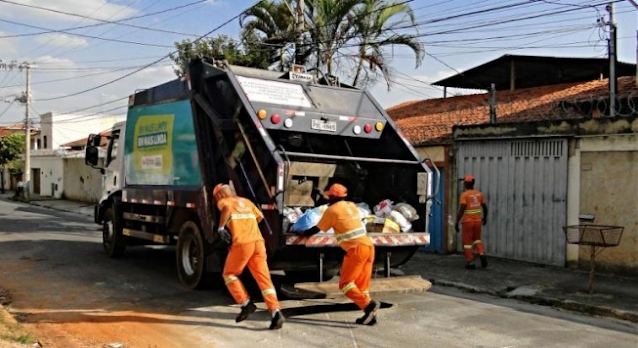 A imagem mostra uma rua residencial, um caminhão de lixo e os catadores de lixo colocando os resíduos dentro do caminhão. Os trabalhadores vestem um uniforme laranja.