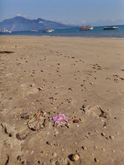 A imagem mostra uma praia. Ao fundo, o mar azul com um morro atrás. Em primeiro plano se vê a areia com máscaras descartadas incorretamente.