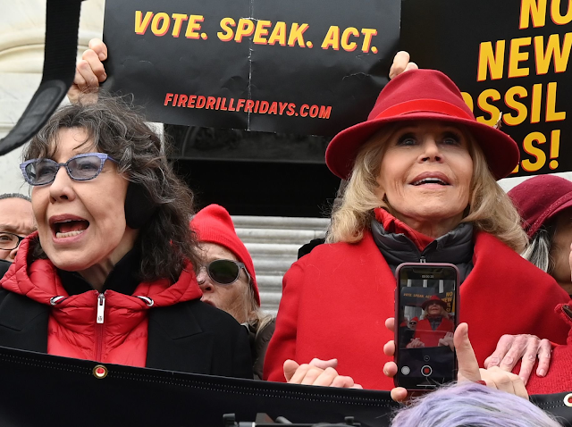 A imagem mostra as atrizes Lily Tomlin e Jane Fonda gritando em um protesto. Elas são brancas e usam casacos de frio.