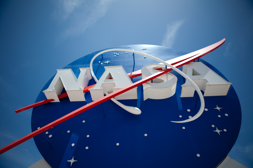 A imagem mostra uma foto do símbolo da NASA. É um círculo azul, com estrelas brancas, escrito NASA em branco com um risco vermelho passando pela palavra.
