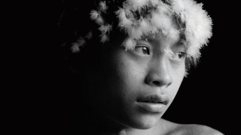 A foto mostra um garoto indígena olhando para o lado. A foto está em preto e branco, com uma leve luz lateral no rosto do garoto. Seus olhos são levemente puxados e sua boca está um pouco aberta. Seu olhar parece que flutua ao longe.