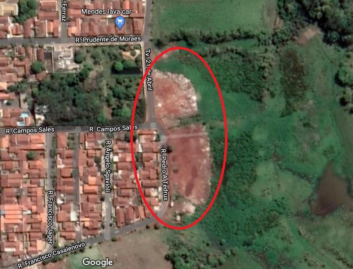 A imagem mostra a área que corresponde ao aterro vista por satélite. A imagem mostra o terreno verde e ao lado as casas em tom marrom. Possui uma pequena área verde entre as casas.